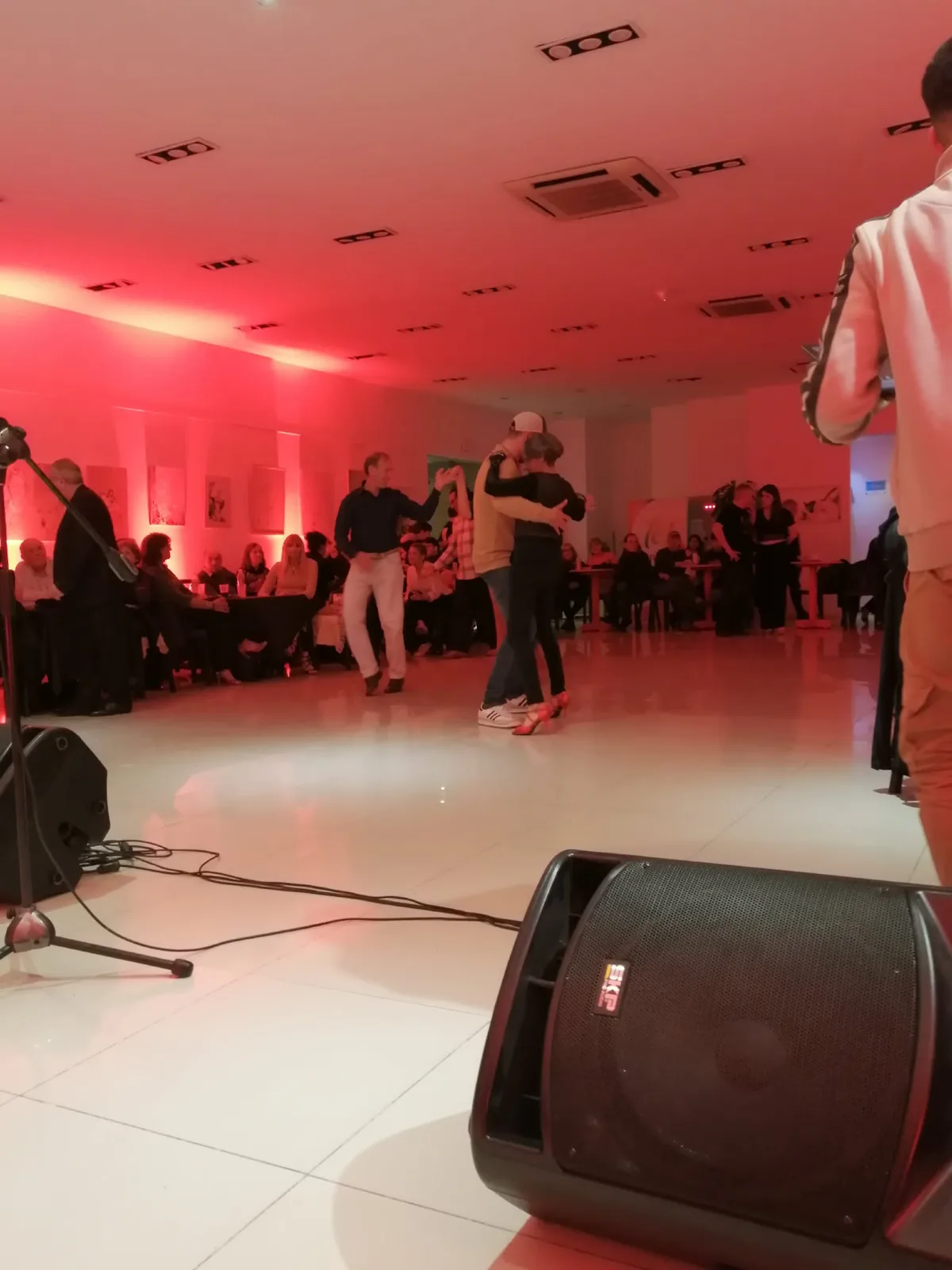 サラテの町のミロンガ。赤い照明の下、白いタイルのフロアで数組が踊り、周囲のテーブルでは客が座って見ている
