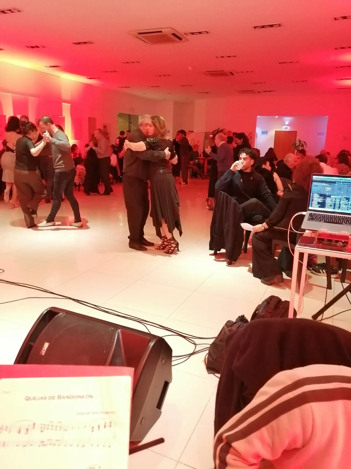同じミロンガの広い画角。奥のフロアで十数組が抱擁して踊り、右手前にはノートパソコンを置いた音響の卓と譜面台がある