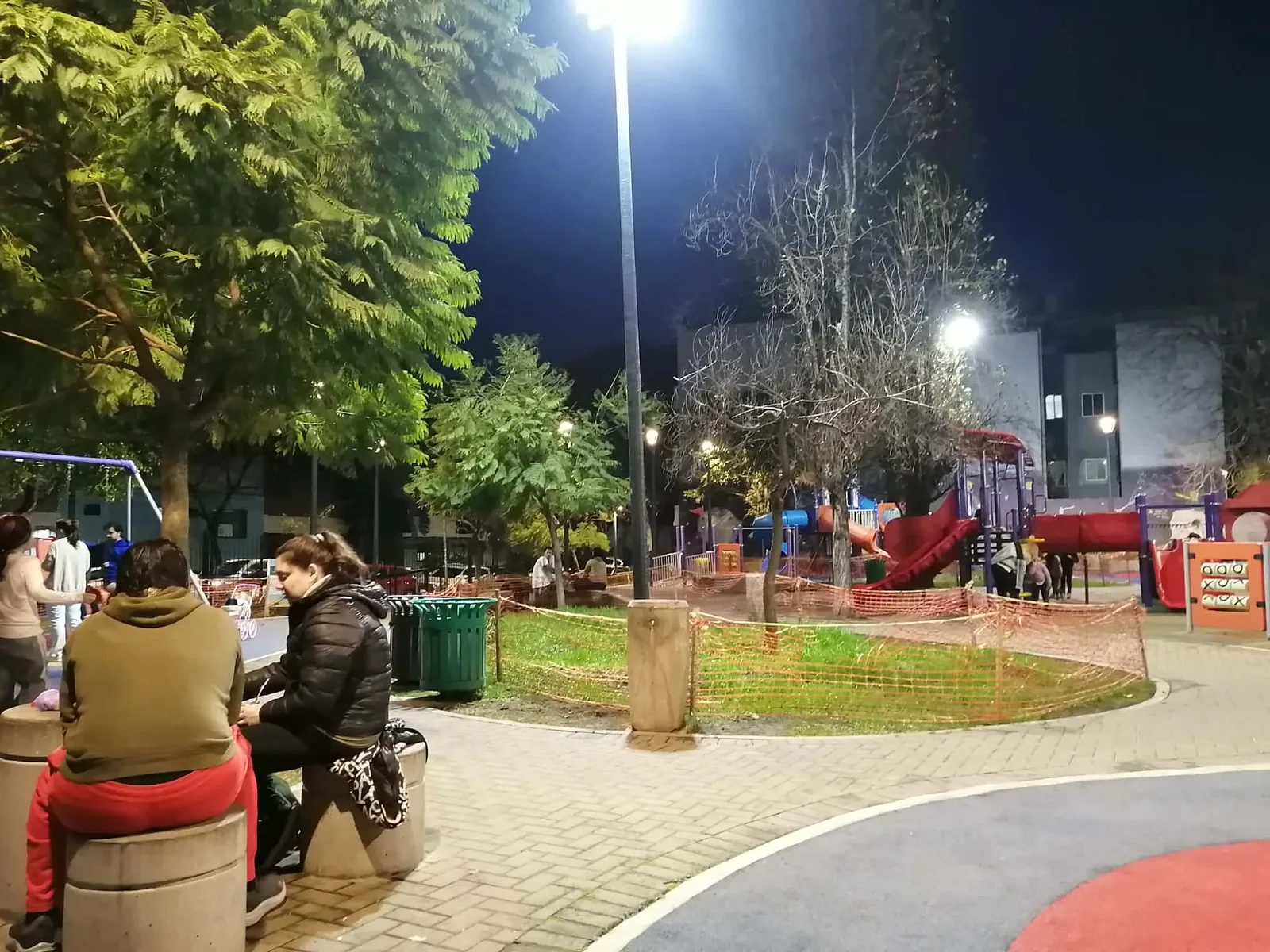 夜の公園。街灯の下で、遊具のまわりに子どもたち、手前のベンチに大人が座っている