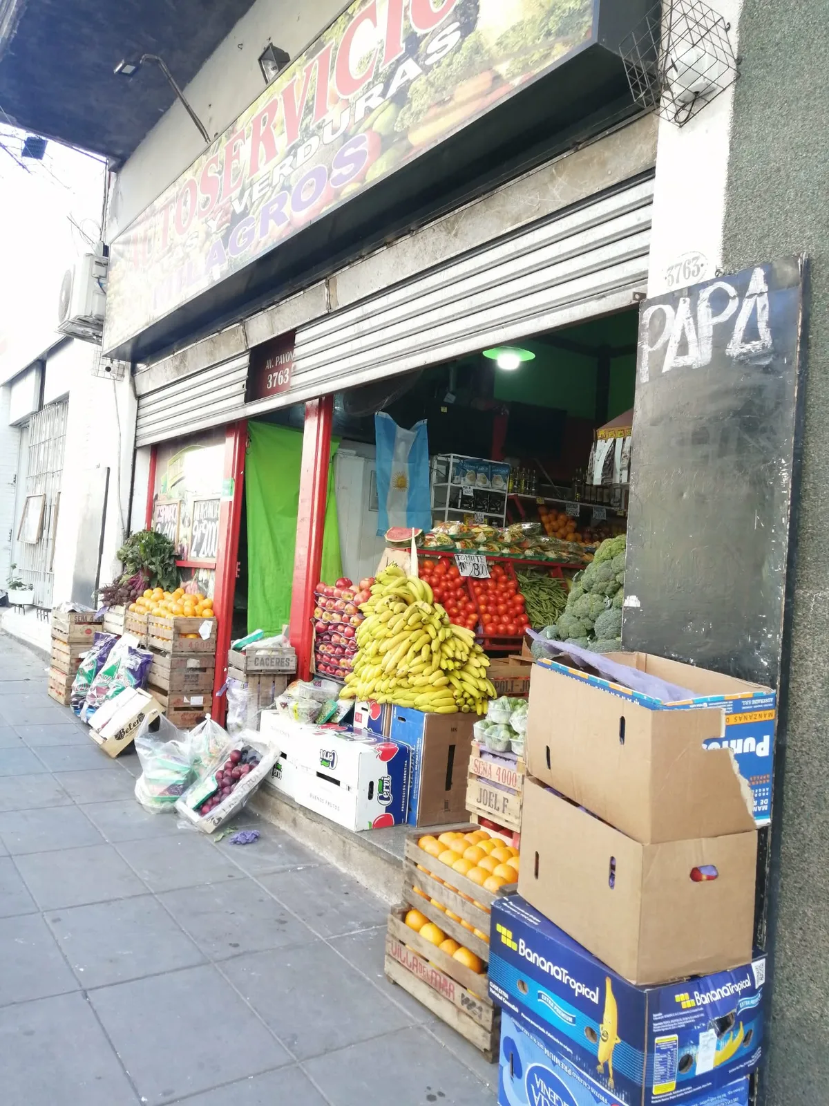 街角の八百屋の店先。歩道まで積まれた果物と野菜の木箱と、店内に掛かるアルゼンチン国旗