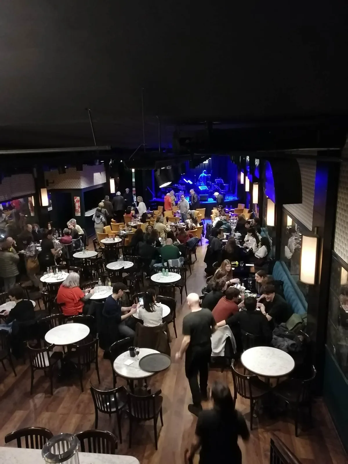 開演前の音楽ホール。丸テーブルの客席がほぼ埋まり、奥の舞台に青い照明が入っている