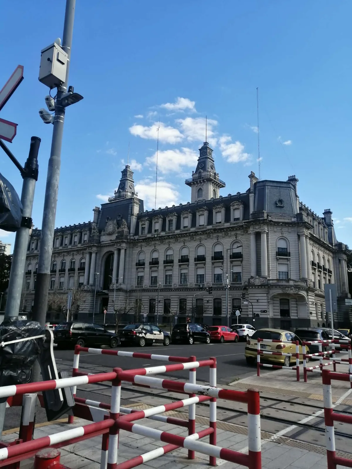 青空の下、塔とスレート葺きの急な屋根を持つ、フランス風の大きな石造りの建物。手前に踏切の赤白の柵