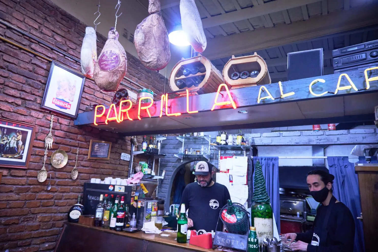 店内。奥にネオンの PARRILLA の文字、天井から生ハムが吊るされ、壁はレンガ積み
