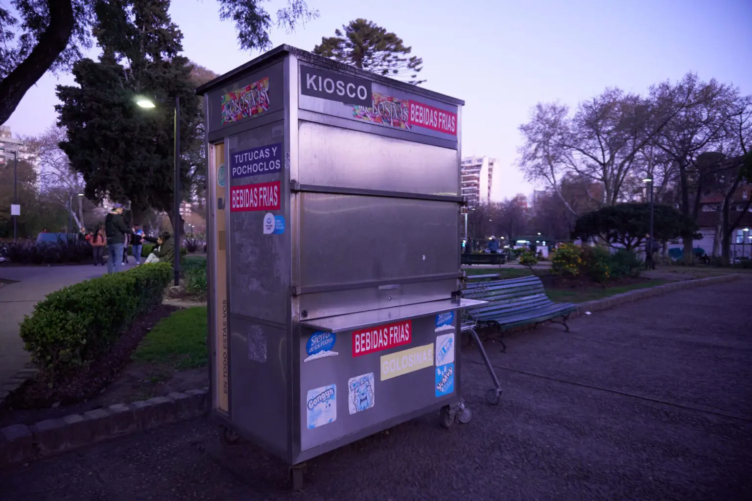 公園に立つ小さなキオスコ。KIOSCO、GOLOSINAS、BEBIDAS FRIAS の看板と、決済アプリのステッカー